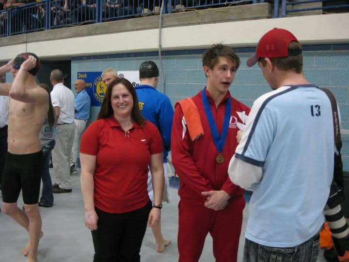 Jon on the pool deck in a Detroit Lakes warmup with a medal around his neck, talking with coaches after a meet.